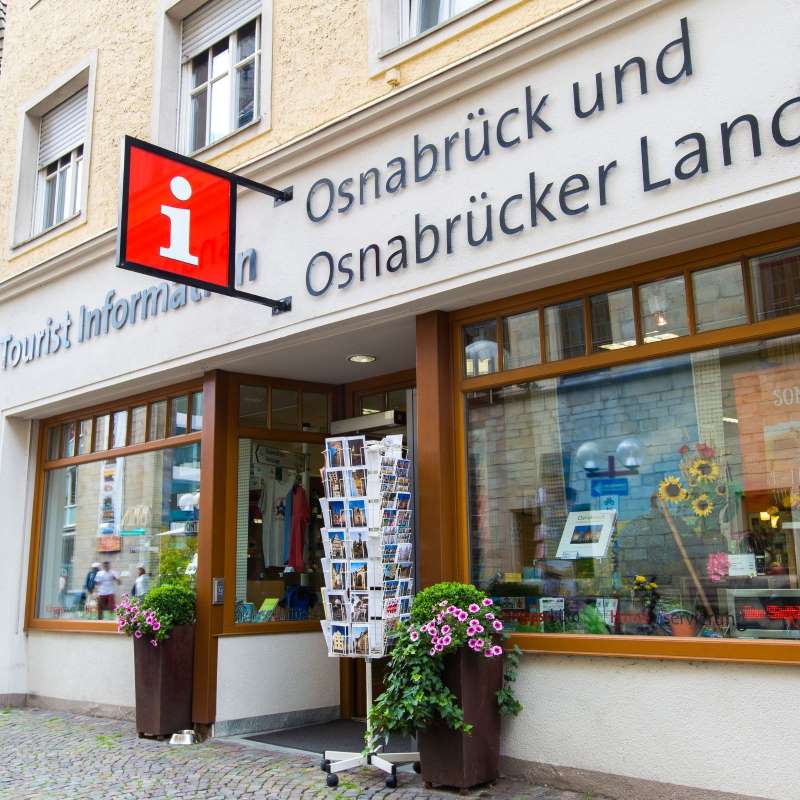 Eingang der Tourist Information mit Postkartenständer, roten Info-Schildern und der Fassadenaufschrift: Osnabrück und Osnabrücker Land.