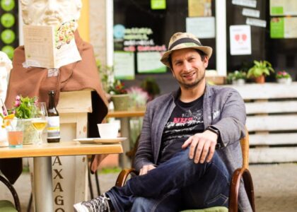 Mann mit Hut sitzt lächelnd vor einem Restaurant im Freien, neben ihm eine Büste mit braunem Tuch und einer Speisekarte am Tisch.
