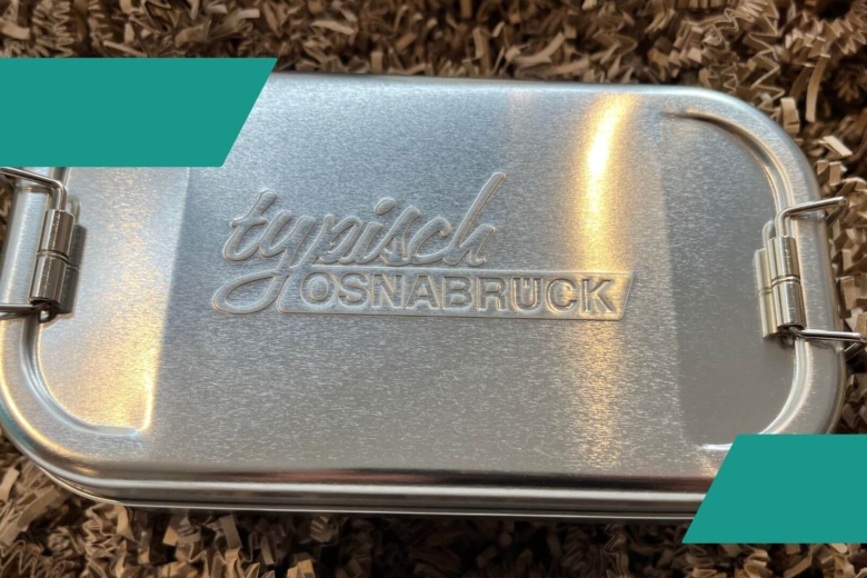 Metallene Lunchbox mit eingeprägtem Schriftzug „Typisch Osnabrück“ auf braunem Füllmaterial.