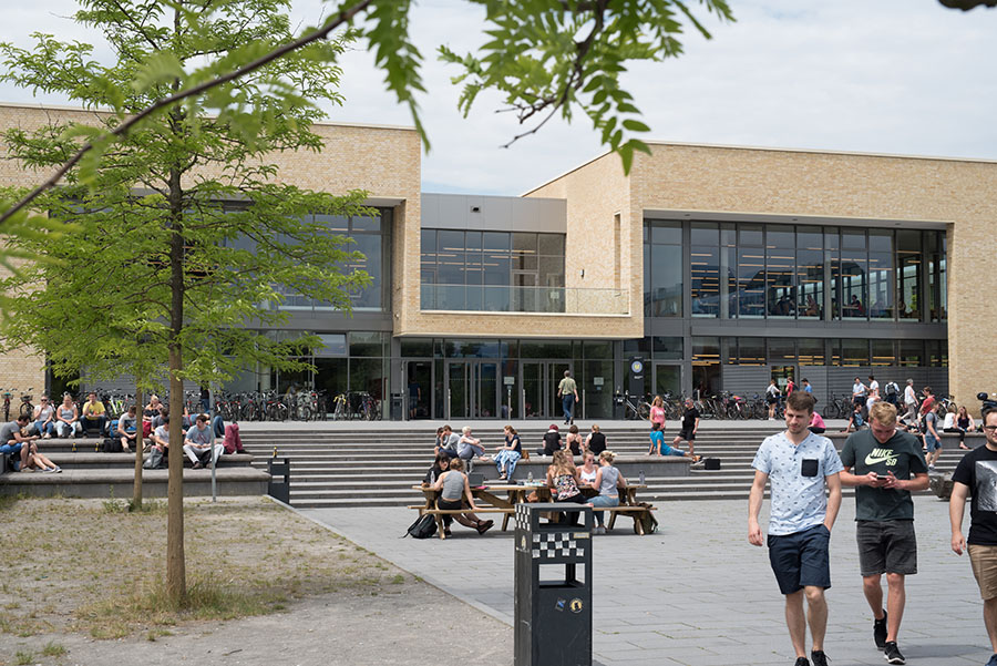 Belebter Campusplatz vor einem modernen Unigebäude mit sitzenden und gehenden Studierenden an Picknicktischen und auf Treppen.