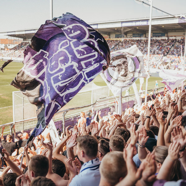 Jubelnde Fußballfans auf der Tribüne eines Stadions schwenken große violette und weiße Fahnen bei einem Spiel.