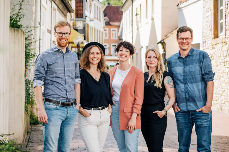Fünf lächelnde Teammitglieder stehen nebeneinander im Freien in einer Altstadtgasse und blicken in die Kamera.