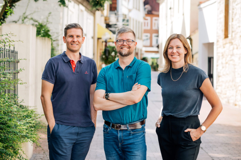 Drei lächelnde Personen, zwei Männer und eine Frau, stehen nebeneinander in einer historischen Altstadtgasse.