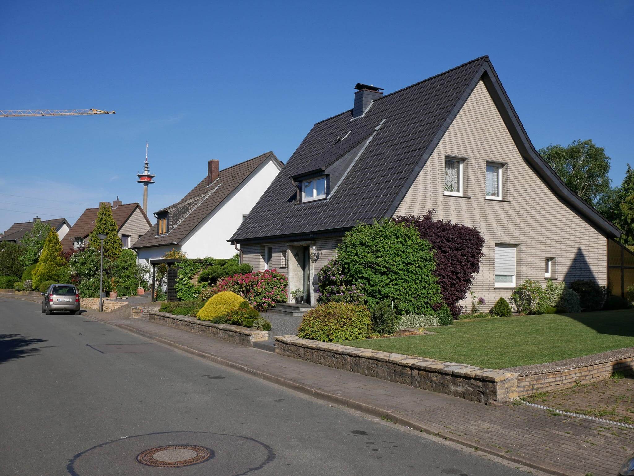 Ruhige Wohnstraße mit Einfamilienhäusern, gepflegten Vorgärten und einem Fernmeldeturm in der Ferne.