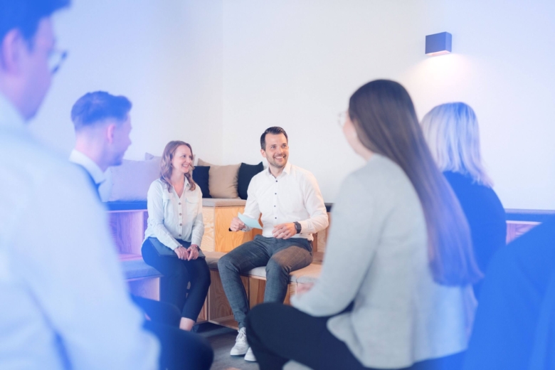Teilnehmende sitzen in einem Workshop im Kreis und hören aufmerksam einem sprechenden Mann in der Mitte zu.
