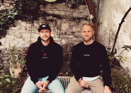 Zwei Männer sitzen nebeneinander auf einer Bank vor einer Steinmauer im Garten, gekleidet in Pullover mit folgendem Schriftzug: Young Diversity.