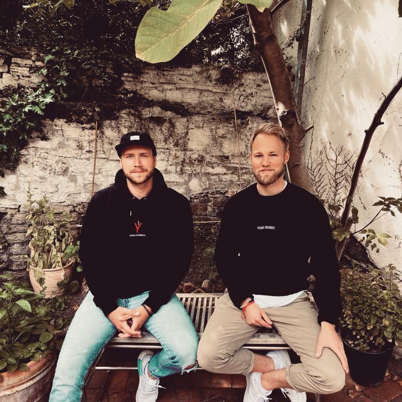 Zwei Männer sitzen nebeneinander auf einer Bank vor einer Steinmauer im Garten, gekleidet in Pullover mit folgendem Schriftzug: Young Diversity.
