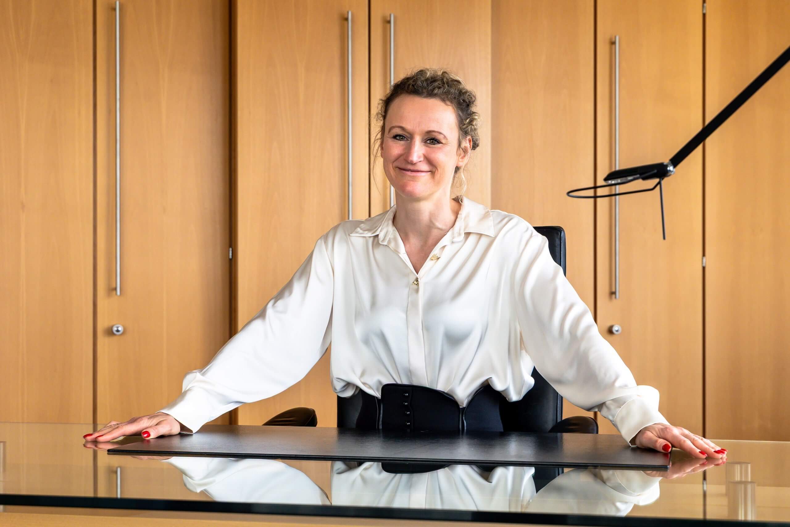 Eine lächelnde Frau mit hochgesteckten Haaren und weißer Bluse stützt sich mit beiden Händen auf ihren Glasschreibtisch im Büro.