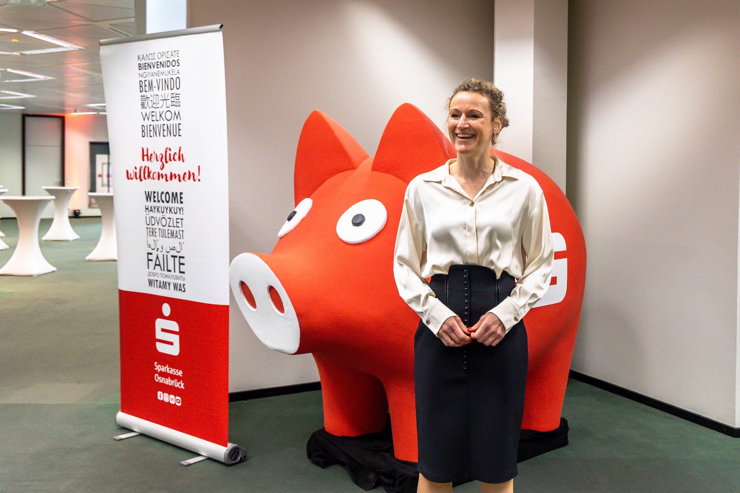Eine lächelnde Frau steht neben einer großen roten Sparkassenschwein-Figur und einem Banner mit der Aufschrift Herzlich willkommen!