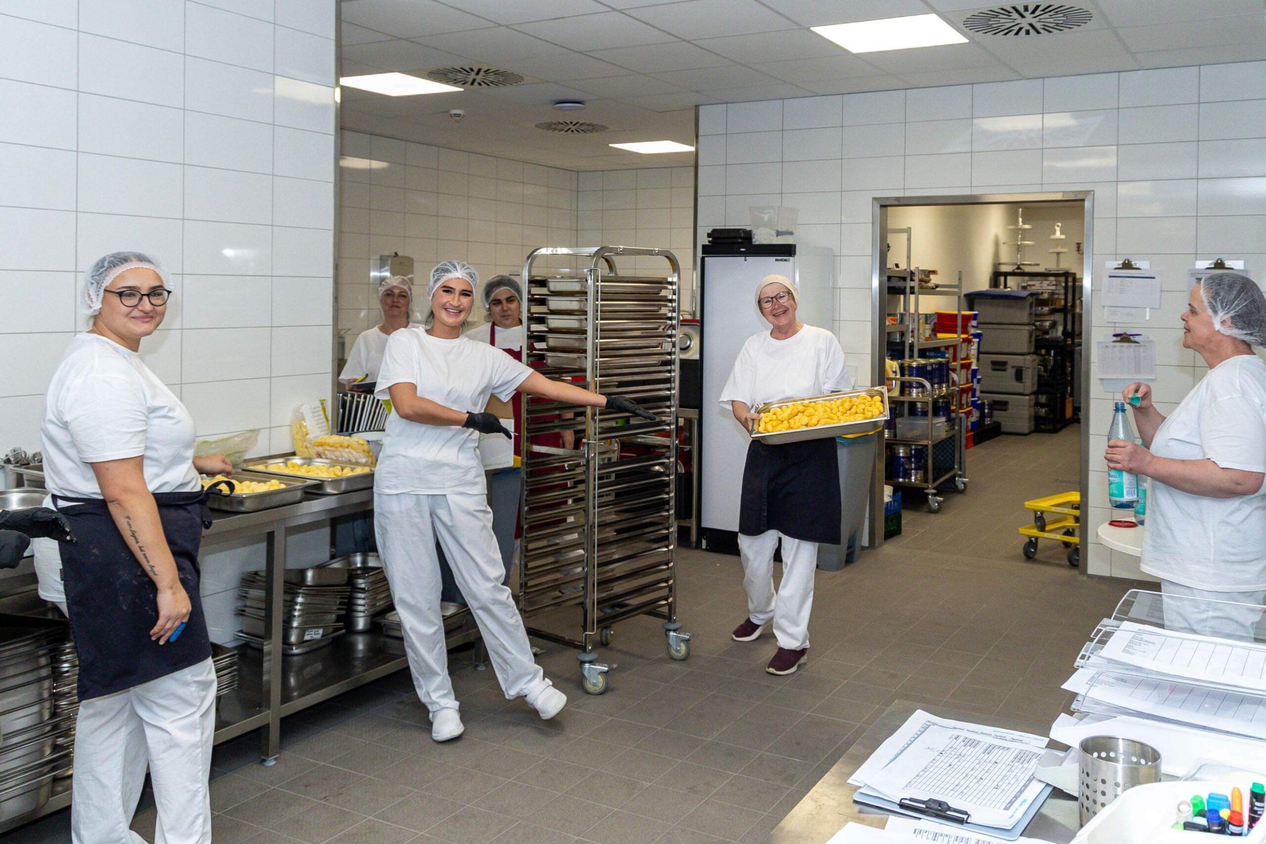 Küchenmitarbeiterinnen in weißer Arbeitskleidung arbeiten lächelnd in einer Großküche.