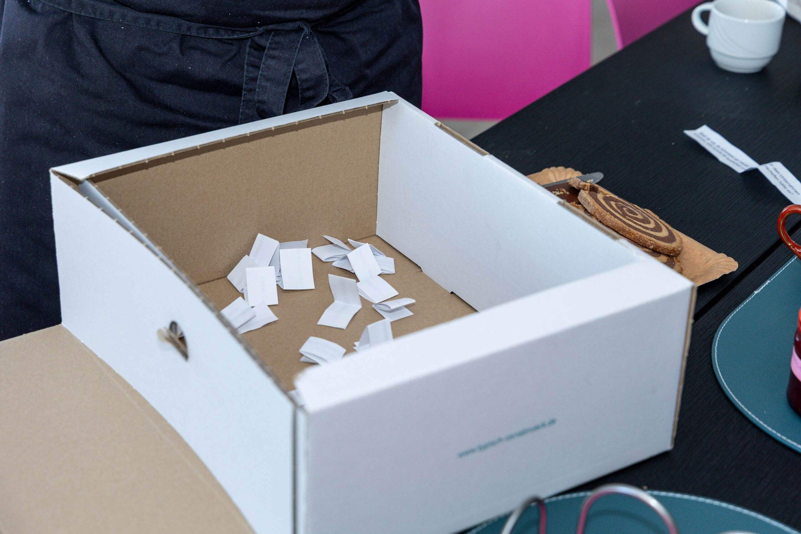Eine offene weiße Kartonkiste mit vielen gefalteten Papierlosen steht auf einem Tisch.