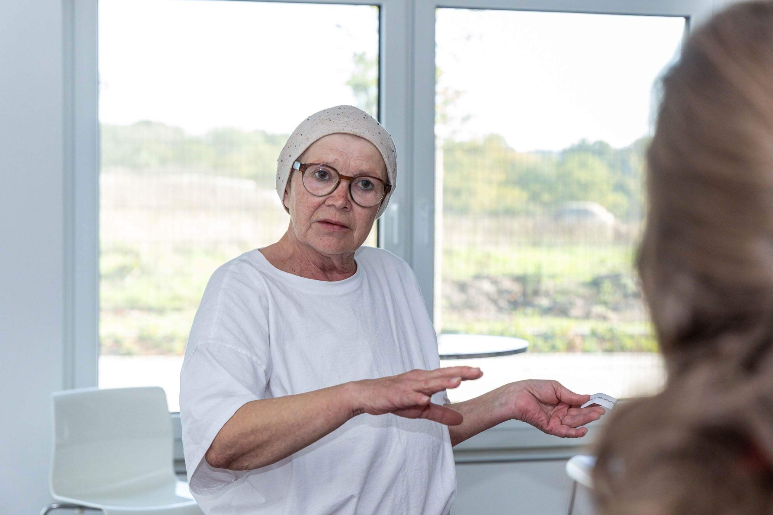 Eine Frau mit Kopftuch und Brille erklärt gestikulierend etwas im Gespräch mit einer Person.