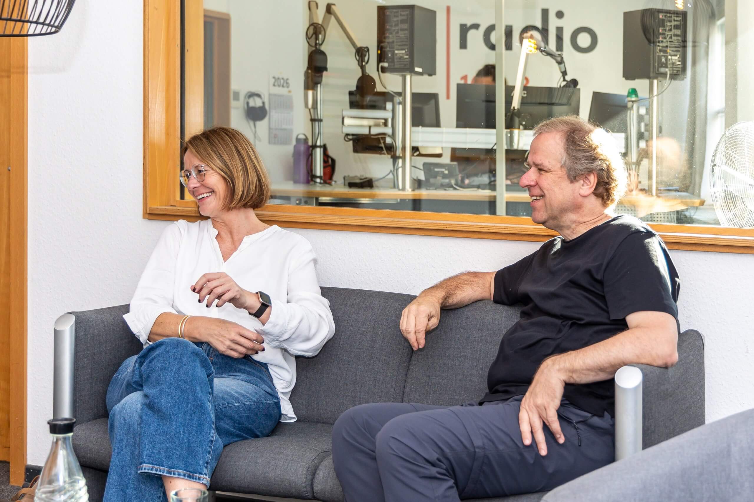 Eine Frau und ein Mann sitzen lachend auf einem grauen Sofa. Im Hintergrund gibt ein Fenster den Blick in ein Radiostudio frei.