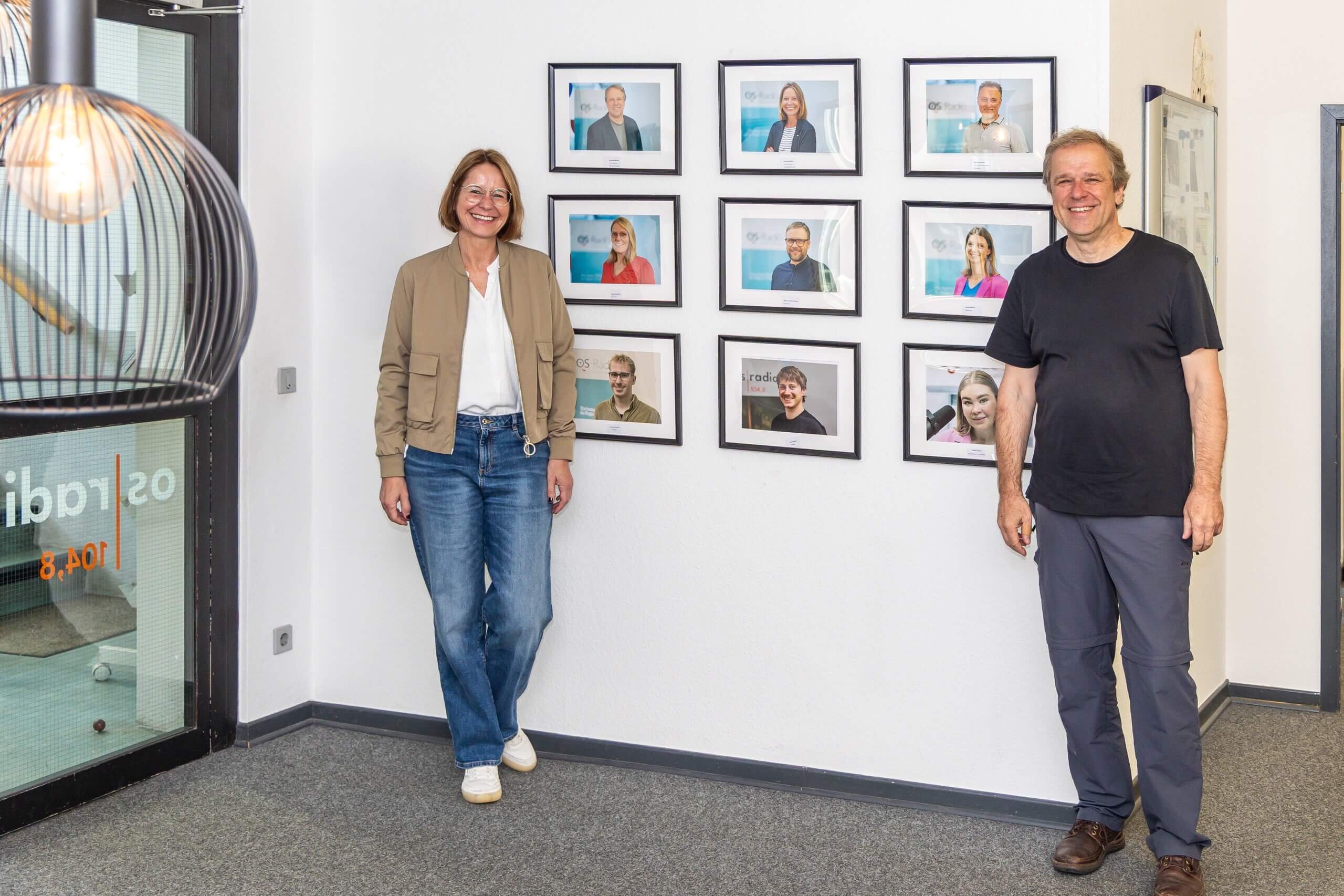 Eine Frau und ein Mann stehen in einem hellen Büro vor einer Wand mit neun gerahmten Portraits. Links ist ein verglaster Eingangsbereich zu sehen.