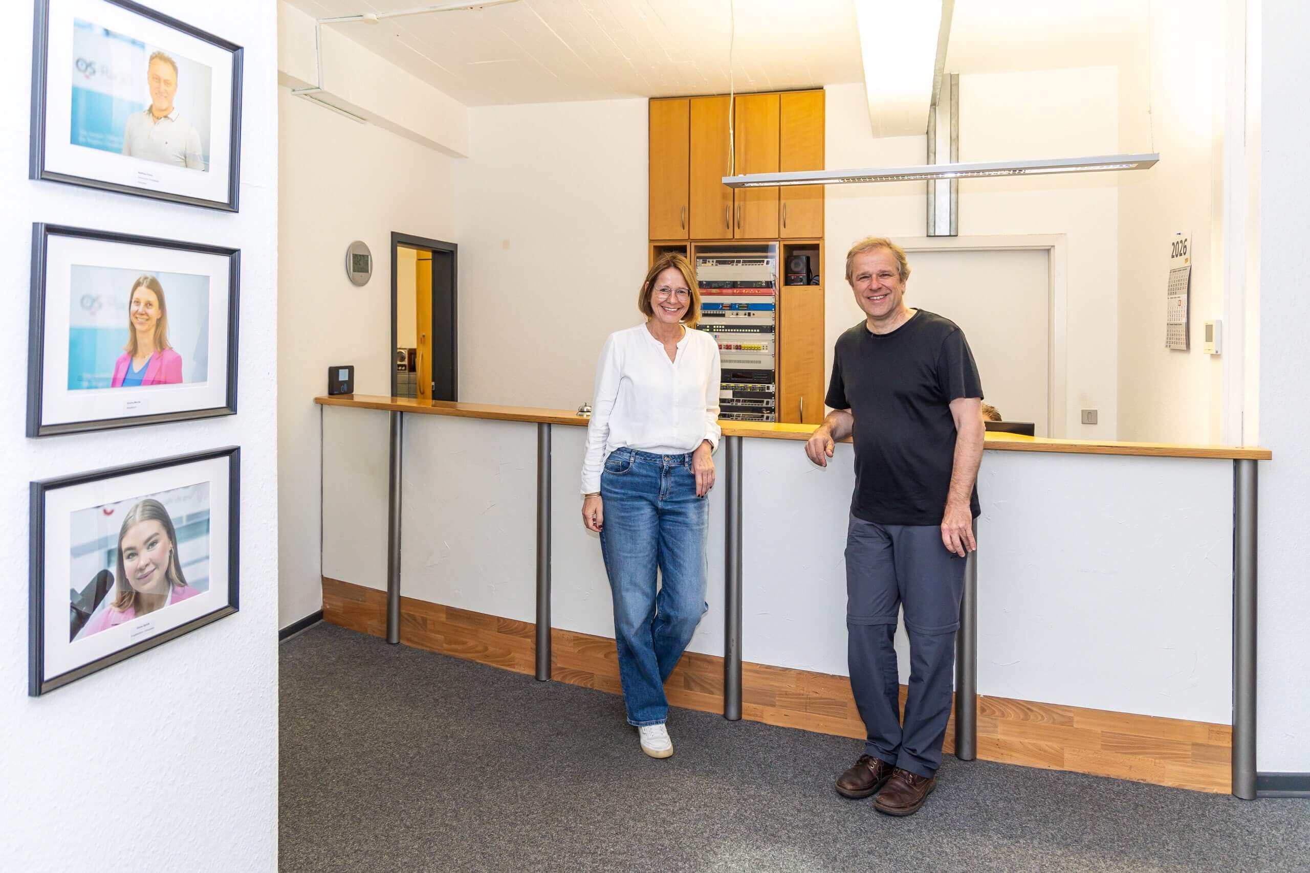 Eine Frau und ein Mann lehnen lächelnd an einem Empfangstresen im Foyer eines Radiostenders. Links hängen gerahmte Porträts an der Wand.