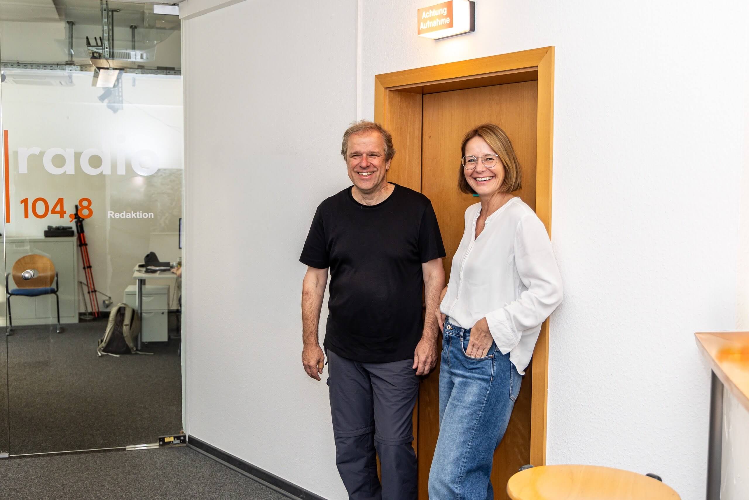 Ein Mann und eine Frau stehen lächelnd im Türrahmen eines Aufnahmeraums bei os radio 104,8. Über ihnen leuchtet das Schild „Achtung Aufnahme“.