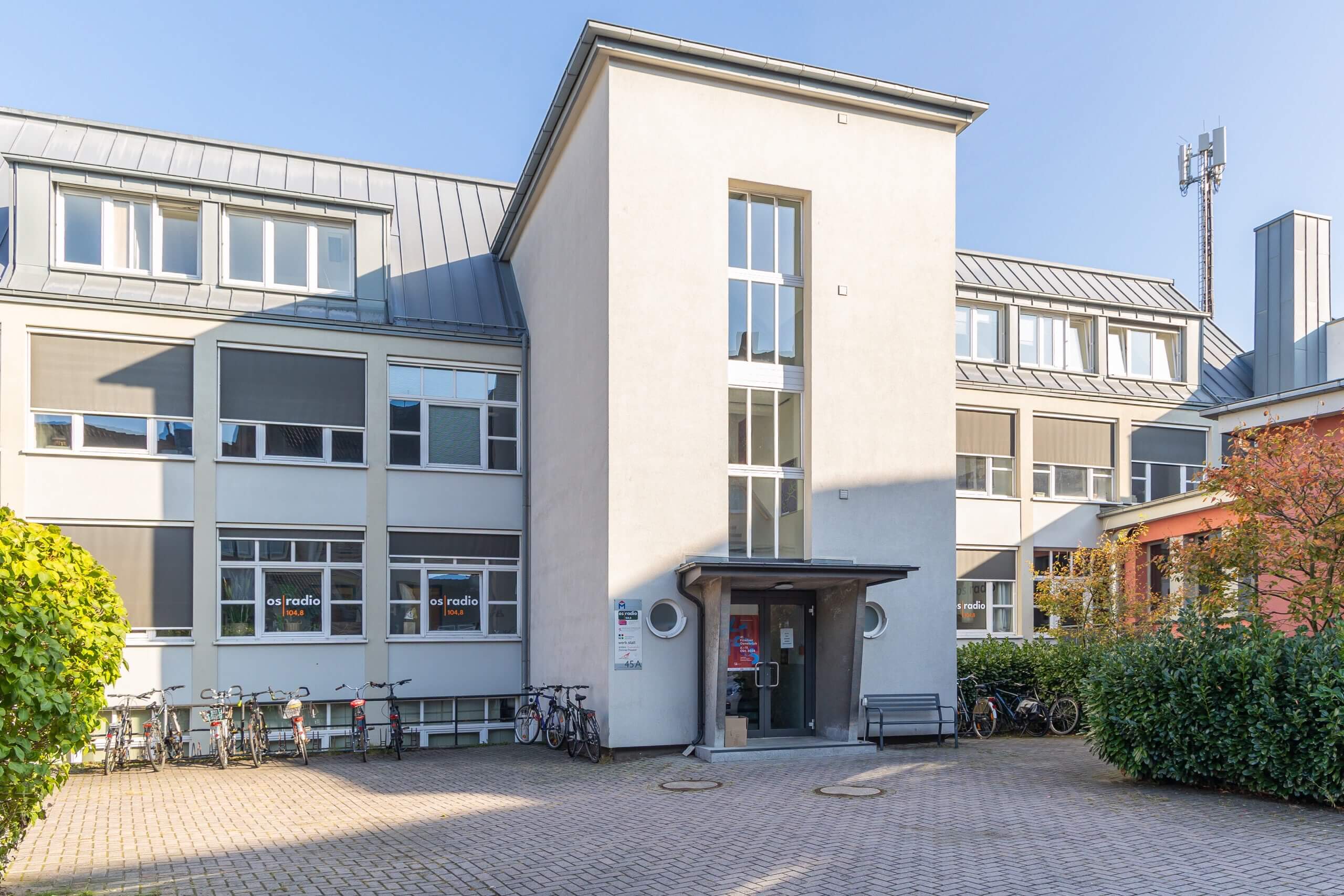 Außenansicht eines mehrstöckigen Gebäudes von os radio 104,8 mit gepflastertem Vorplatz, geparkten Fahrrädern und Eingangsbereich.