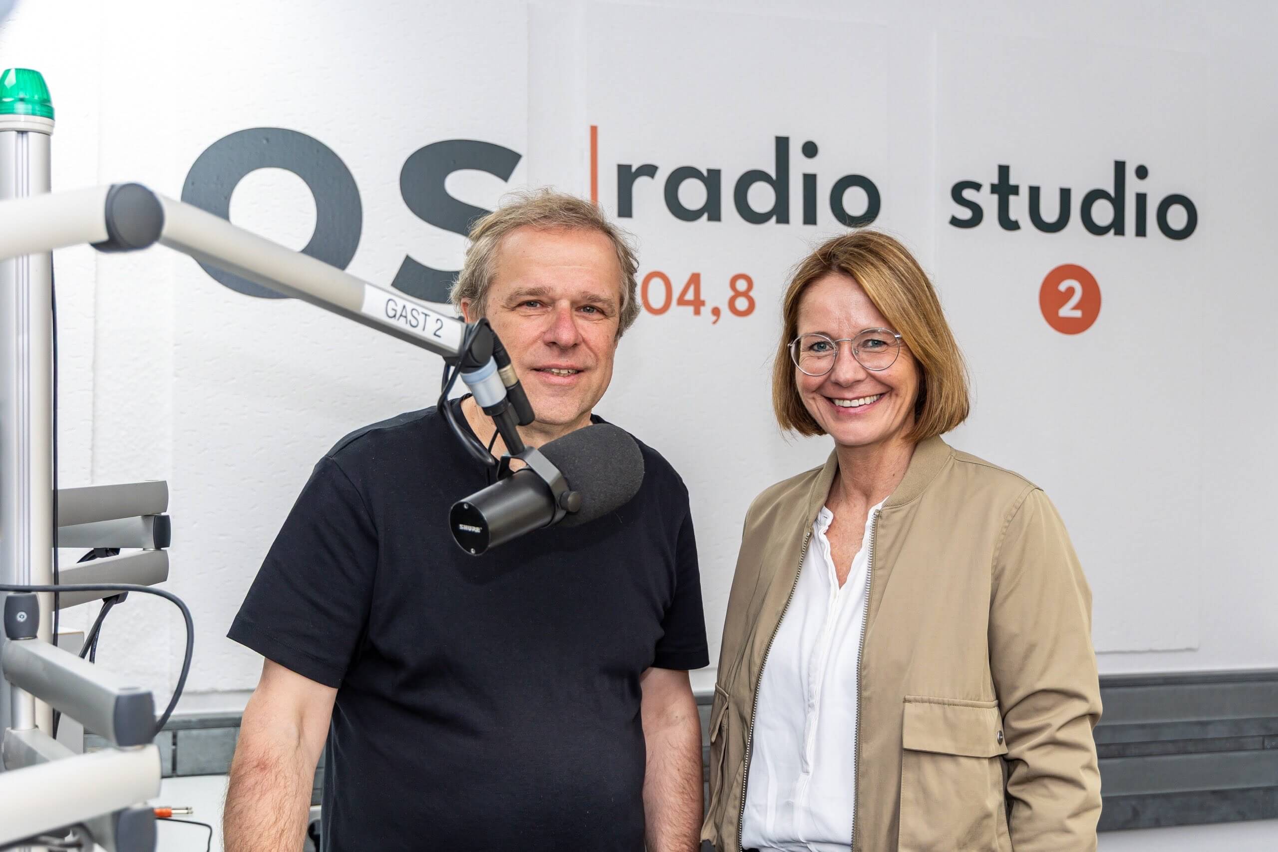 Ein Mann und eine Frau lächeln im Studio von os radio 104,8 nebeneinander in die Kamera. Im Vordergrund steht ein Studiomikrofon.