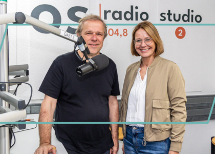 Ein Mann und eine Frau lächeln im Studio von os radio 104,8 nebeneinander in die Kamera. Im Vordergrund steht ein Studiomikrofon.