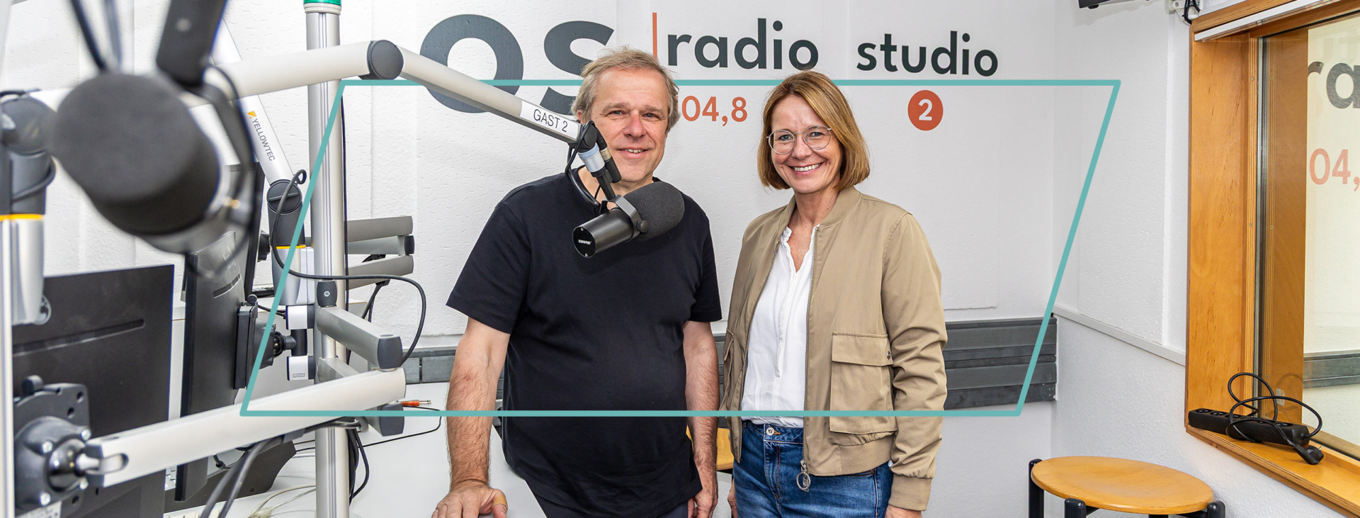 Ein Mann und eine Frau lächeln im Studio von os radio 104,8 nebeneinander in die Kamera. Im Vordergrund steht ein Studiomikrofon.